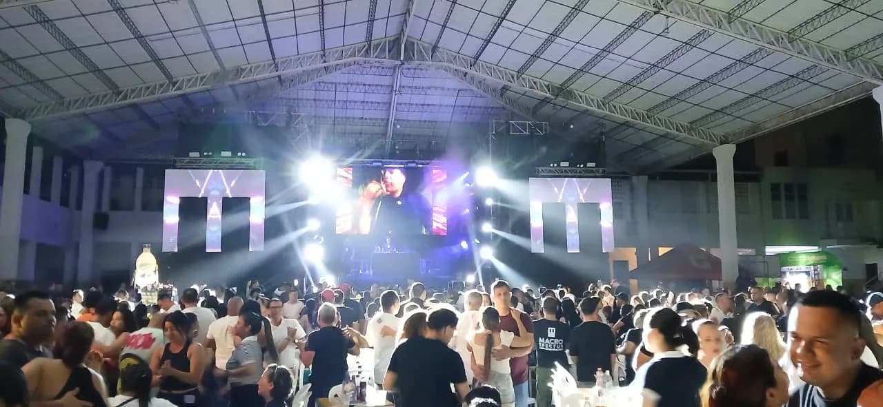 Macro Efectus en tarima ante un coliseo techado lleno: las pantallas LED con la «M» estilizada de la agrupación a cada lado del escenario, los cañones de luz abriéndose sobre el público y, entre la gente, un asistente con la camiseta de la casa
