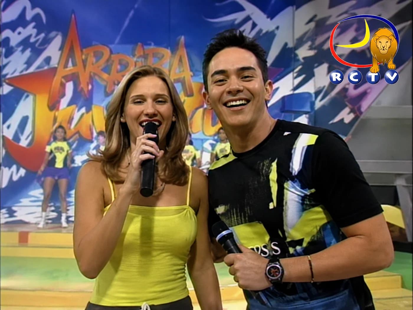 Camila Canabal, microphone in hand, beside the other host of “Arriba Juventud” on the set of the RCTV show, with the show's sign at the back and the channel's logo on screen