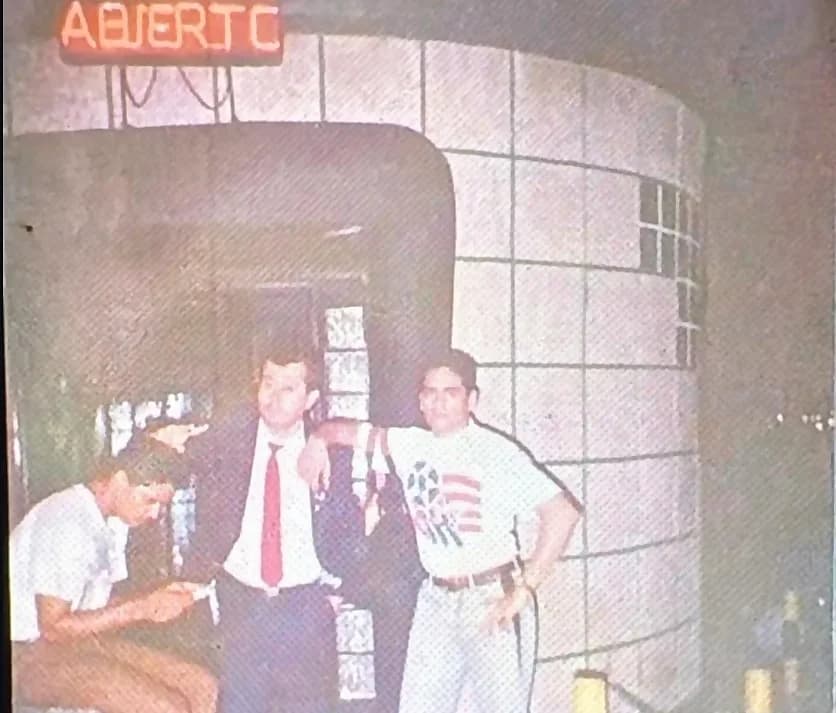 The entrance to the Glass Club at night: the curved tiled façade with glass blocks and the red neon sign “ABIERTO” lit above the door, with three men talking in the doorway