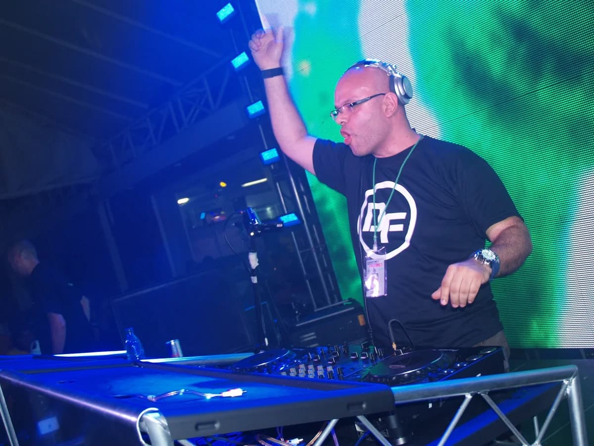 Daniel Frontado en cabina al aire libre, de lentes y camiseta negra con el logotipo «DF», con un brazo en alto tras la mesa de mezclas