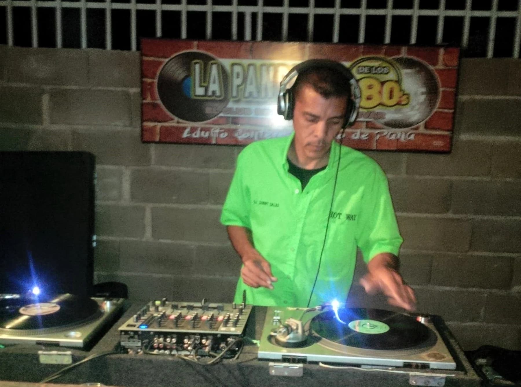 Danny Salas mezclando en dos tornamesas de vinilo, con audífonos y la camisa verde de la miniteca bordada «DJ DANNY SALAS» en el pecho izquierdo y «HOT WAY» en el derecho, bajo un letrero de club con discos de vinilo y la leyenda «de los 80s»