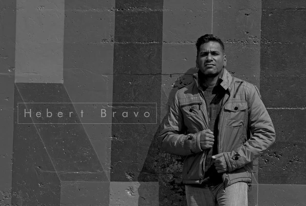 Hebert Bravo en retrato blanco y negro de cuerpo entero, con chaqueta, ante una pared de formas geométricas con su nombre rotulado