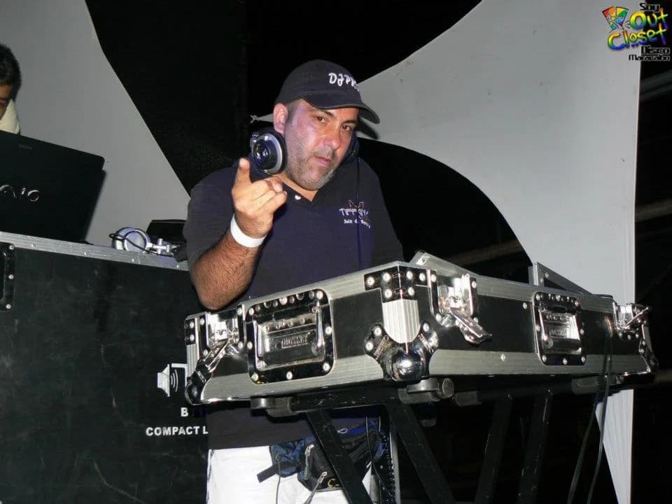 DJ Pro Venezuela con gorra y camisa oscura tras una hilera de flight cases metálicos, sosteniendo un audífono en la mano, con una pancarta de colores al fondo