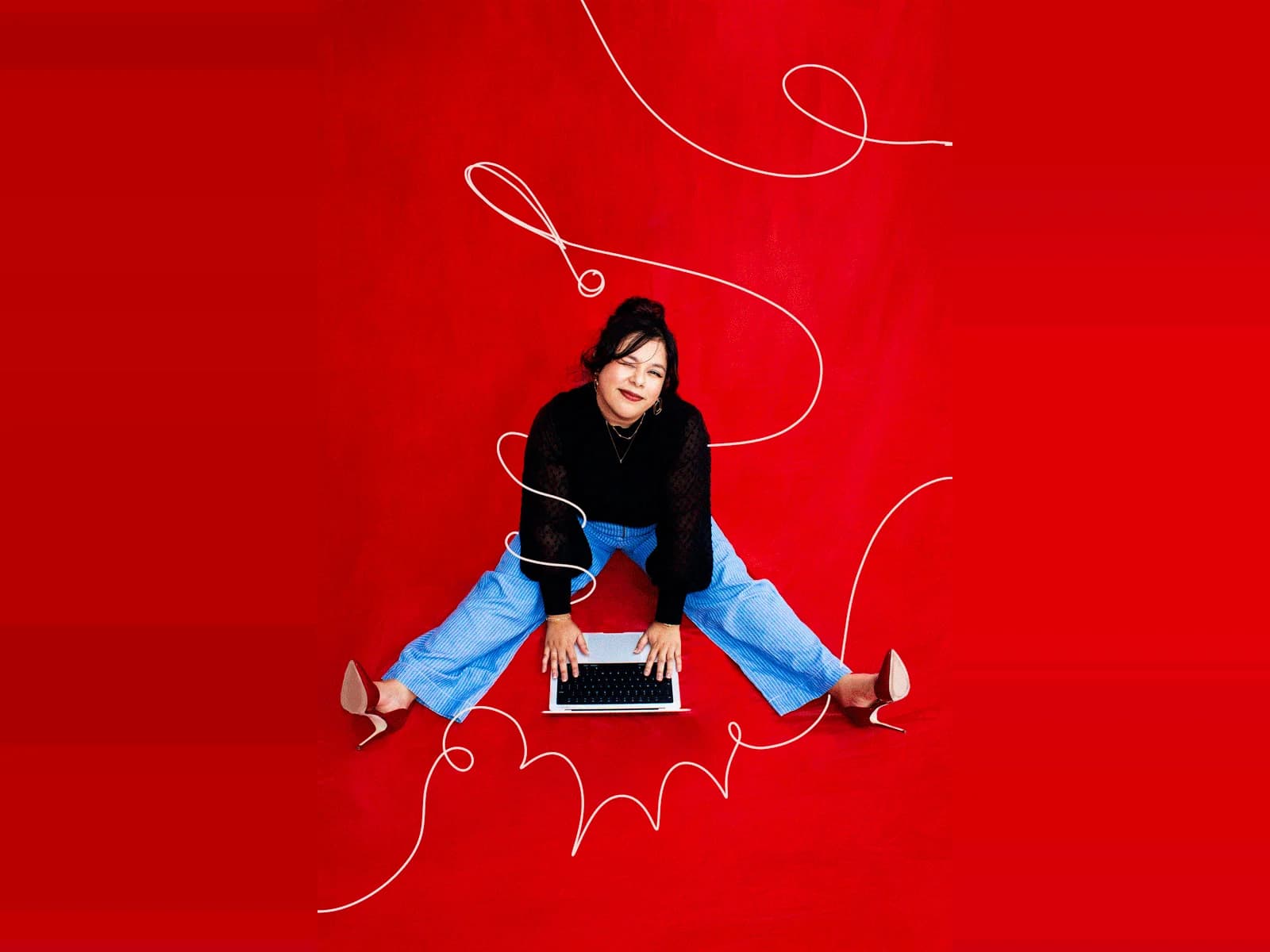 Vicky Rojas-Smith, founder of Alma Co., sitting on the floor at a laptop against a red backdrop crossed by a single continuous white line — studio portrait