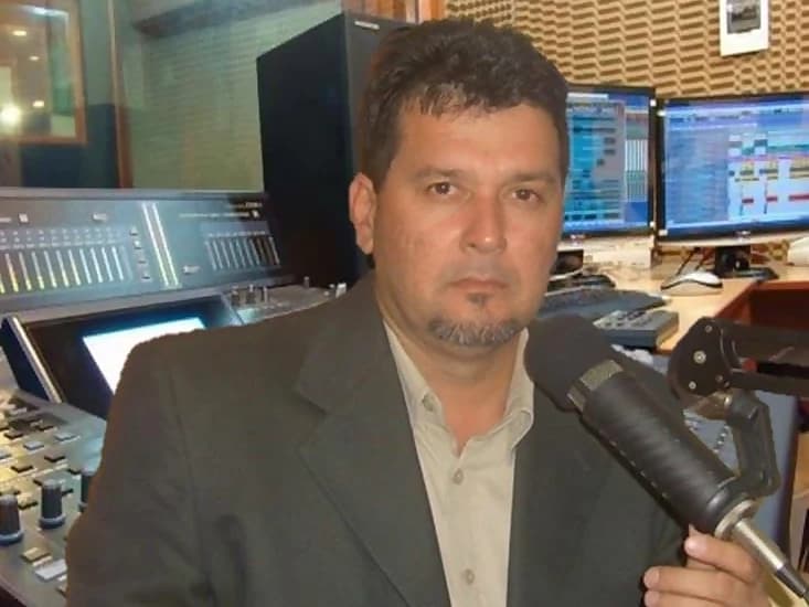 Henry White at the radio booth microphone, in a dark suit, with the console and the programming screens behind him