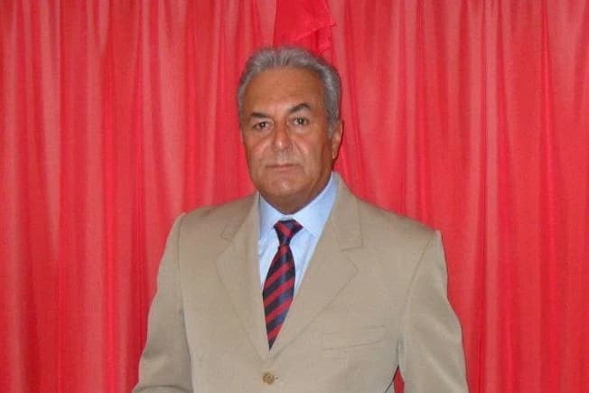 Retrato del locutor José Luis Simoza, de traje claro y corbata a rayas, ante un fondo de cortinas rojas