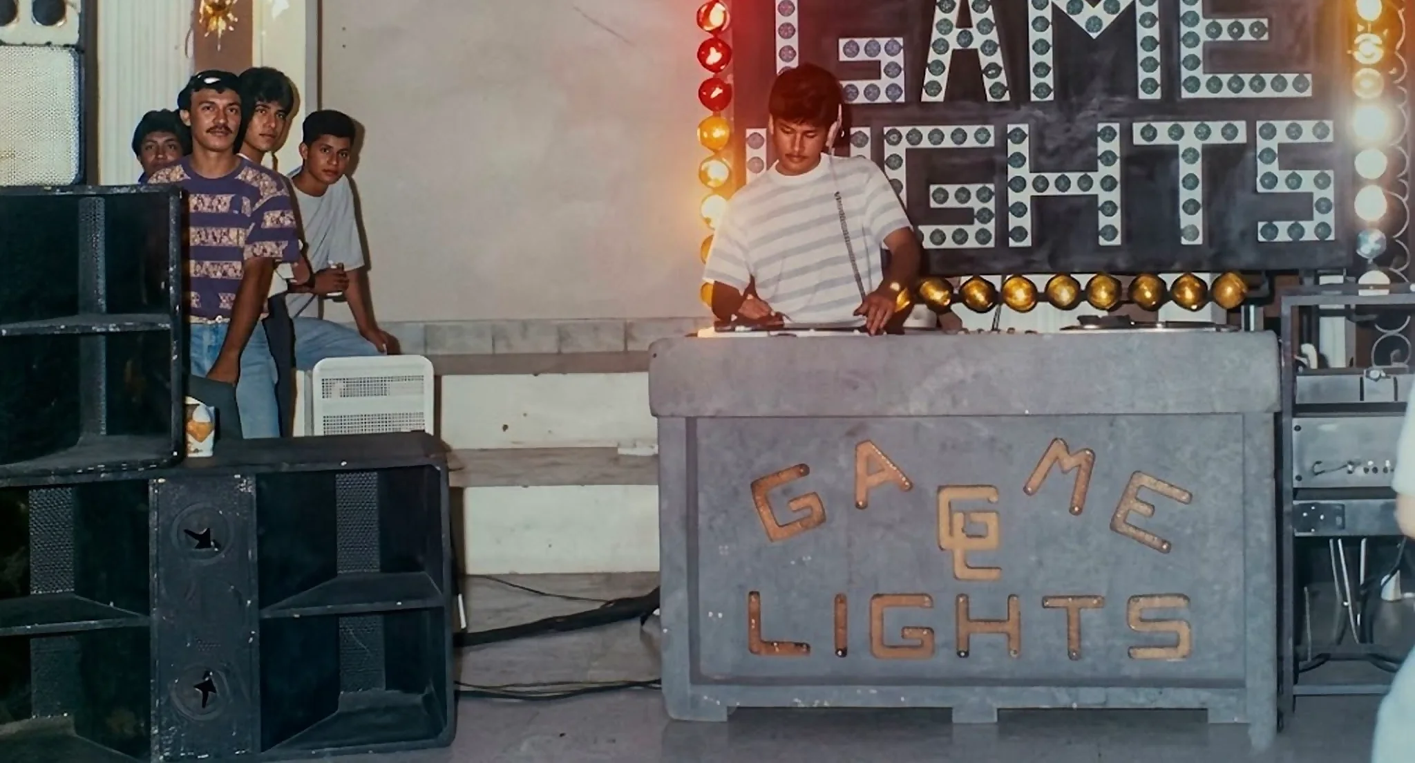 Miniteca Game Lights — el discjockey en la cabina bajo el letrero luminoso «Game Lights», con un grupo de jóvenes junto al equipo, fotografía de archivo