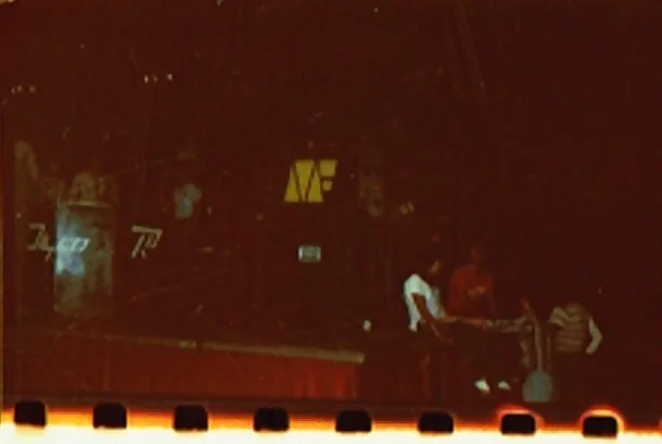 Miniteca Music Factory — fotografía de archivo de la tarima a oscuras con el logo «MF» iluminado en amarillo al centro de la cabina y parte del equipo sentado al borde del escenario (4ª Guerra del Poliedro, 1980)