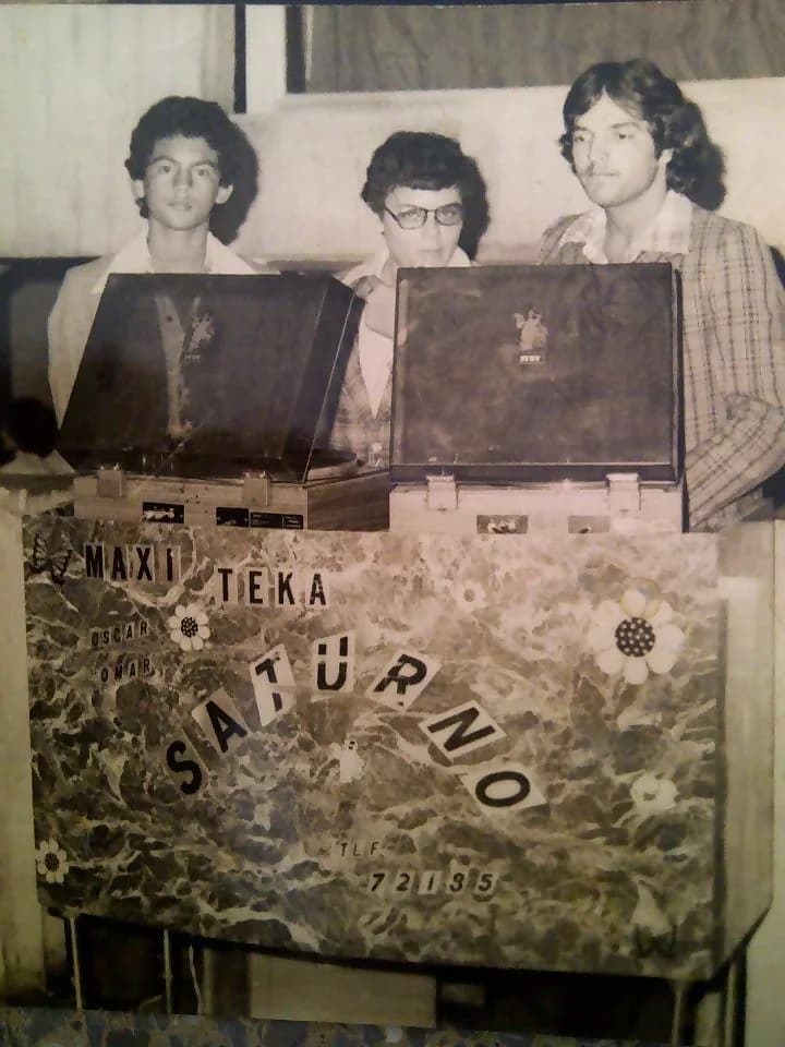 Miniteca Saturno — three young crew members behind the two turntables of the marbled cabinet lettered “Maxi Teka Saturno”, with the names Oscar and Omar and the period telephone number, photograph from the 1970s