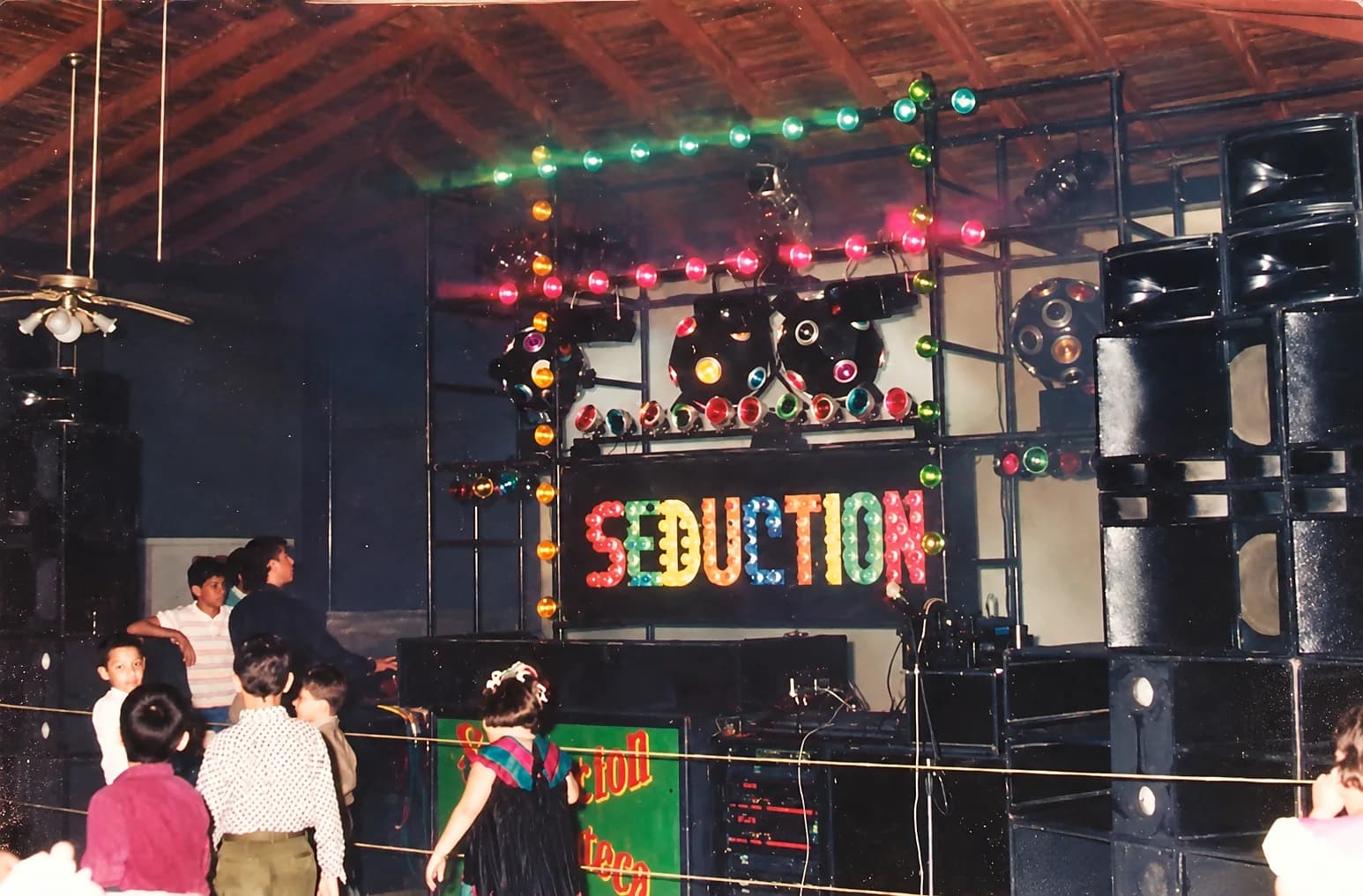 Miniteca Seduction — el letrero de bombillos multicolor «Seduction» sobre la cabina, con niños bailando frente a la tarima, fotografía de archivo