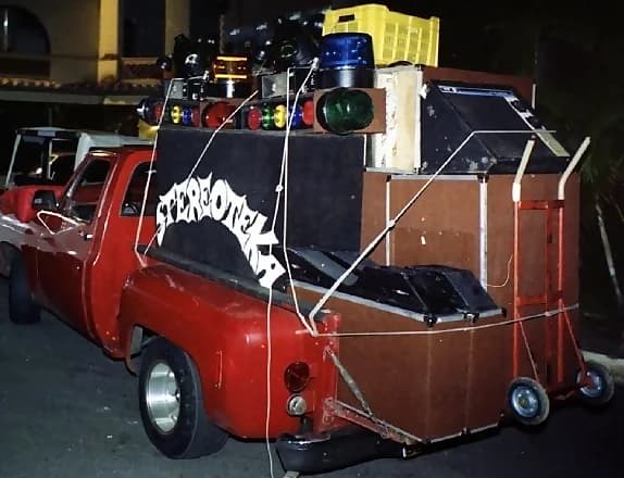 Transporte de la miniteca Stereoteka — camioneta roja con el mueble de cabina rotulado «STEREOTEKA», cargado con las luces y equipos del sistema