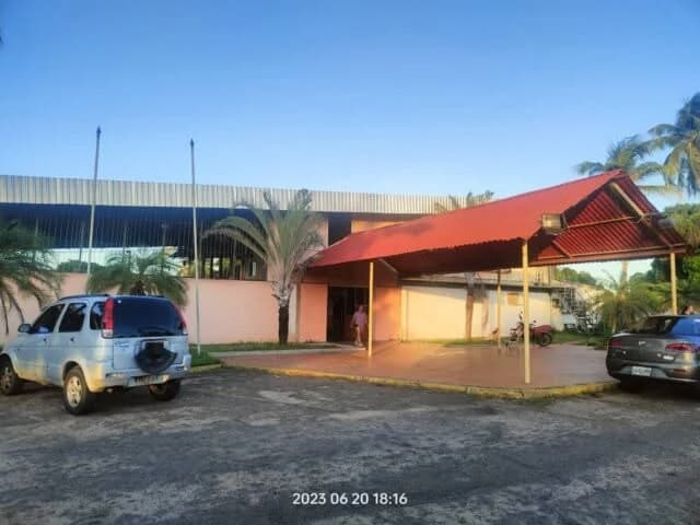 The premises of the Club de Comercio de Ciudad Bolívar seen from the parking lot: the low body of the hall with its metal roof, the entrance porch with its red gabled cover, the palms of the garden and the flagpoles out front