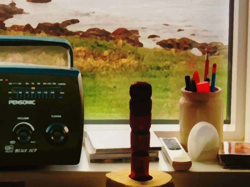 A vintage portable radio in the window, pencils and papers beside it and the green of the coast behind: Saturday morning — cover art of the CMTK Radio show Clásicos de la Mañana