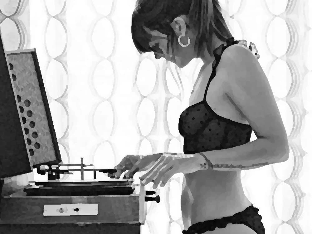 Black-and-white photograph: a young woman lowers the needle of a suitcase record player beside the curtain — cover art of the CMTK Radio show Mañanas de Colección