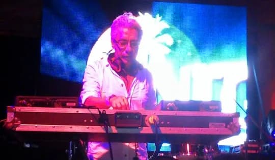 DJ Qchi en cabina, de camisa clara, mezclando sobre reproductores montados en flight case ante una pantalla de video azul DJ Qchi en cabina, de camisa clara, mezclando sobre reproductores montados en flight case ante una pantalla de video azul
