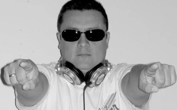 DJ Tille en retrato blanco y negro, con lentes de sol y audífonos al cuello, señalando con ambas manos hacia la cámara DJ Tille en retrato blanco y negro, con lentes de sol y audífonos al cuello, señalando con ambas manos hacia la cámara