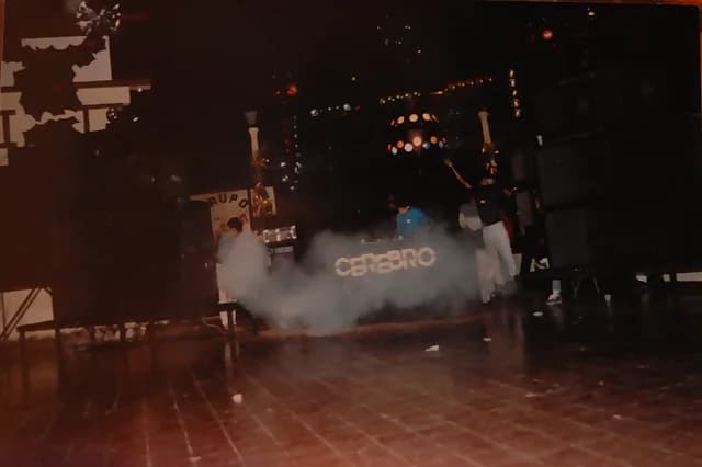 Miniteca Cerebro — the booth bearing the name “Cerebro” emerging through the smoke on the dance floor, with the light sphere lit and the crew working at a period party Miniteca Cerebro — the booth bearing the name “Cerebro” emerging through the smoke on the dance floor, with the light sphere lit and the crew working at a period party
