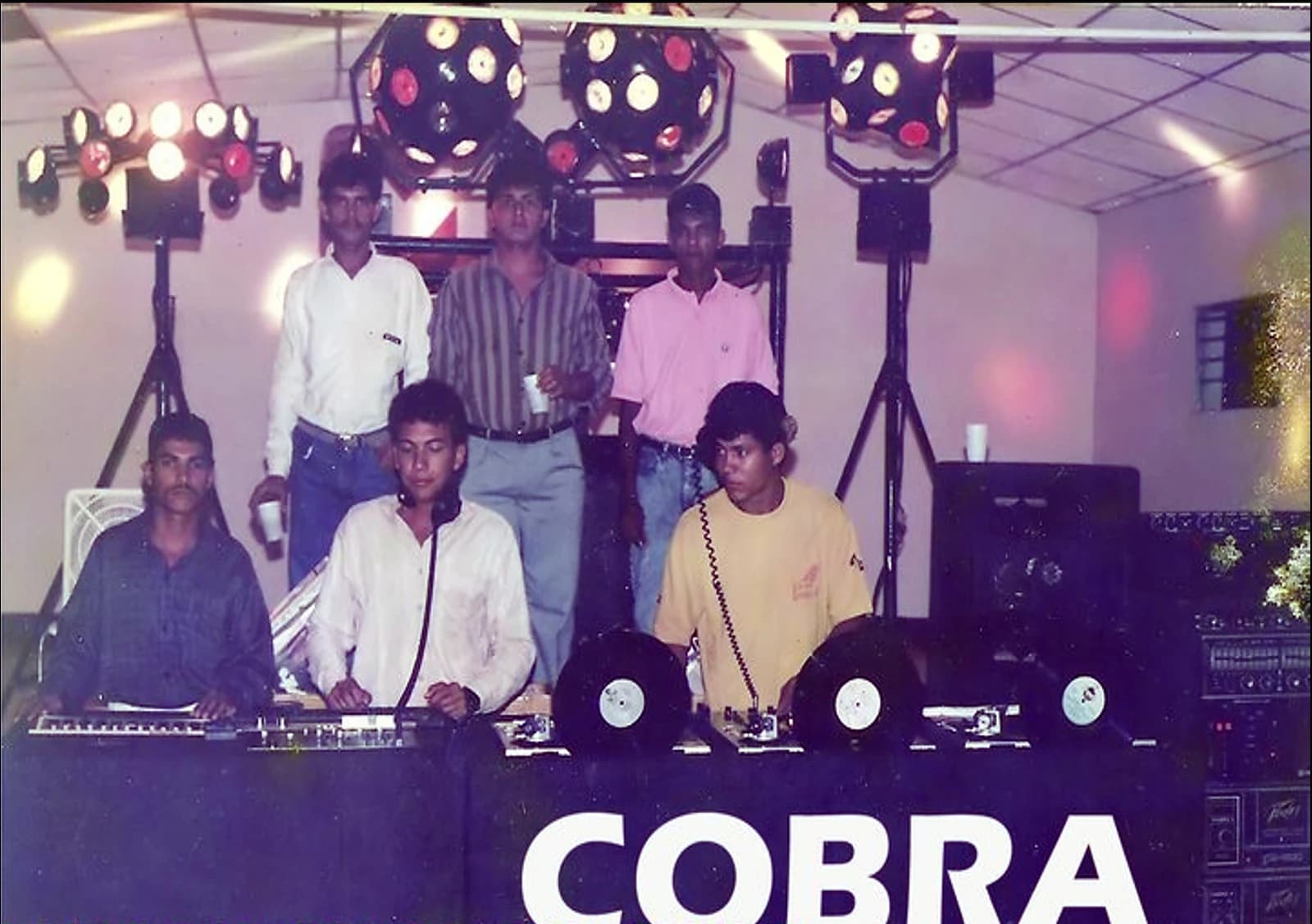 Miniteca Cobra — el equipo de discjockeys en la cabina rotulada «COBRA», con los platos y los discos de vinilo al frente, las esferas de luces con discos girando en lo alto y las torres de sonido a un lado Miniteca Cobra — el equipo de discjockeys en la cabina rotulada «COBRA», con los platos y los discos de vinilo al frente, las esferas de luces con discos girando en lo alto y las torres de sonido a un lado