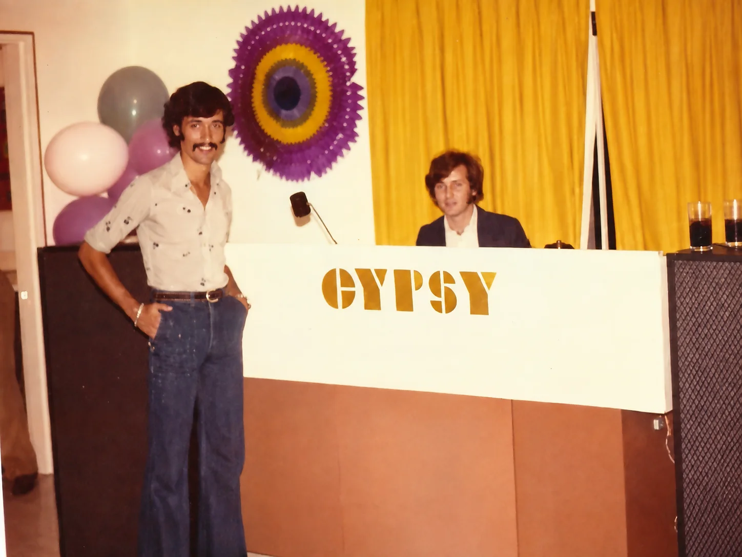 Miniteca Gypsy — dos integrantes junto al mueble blanco rotulado «GYPSY» en letras doradas, con las cornetas a los lados y globos y un abanico de papel al fondo, en una fiesta de los años setenta Miniteca Gypsy — dos integrantes junto al mueble blanco rotulado «GYPSY» en letras doradas, con las cornetas a los lados y globos y un abanico de papel al fondo, en una fiesta de los años setenta