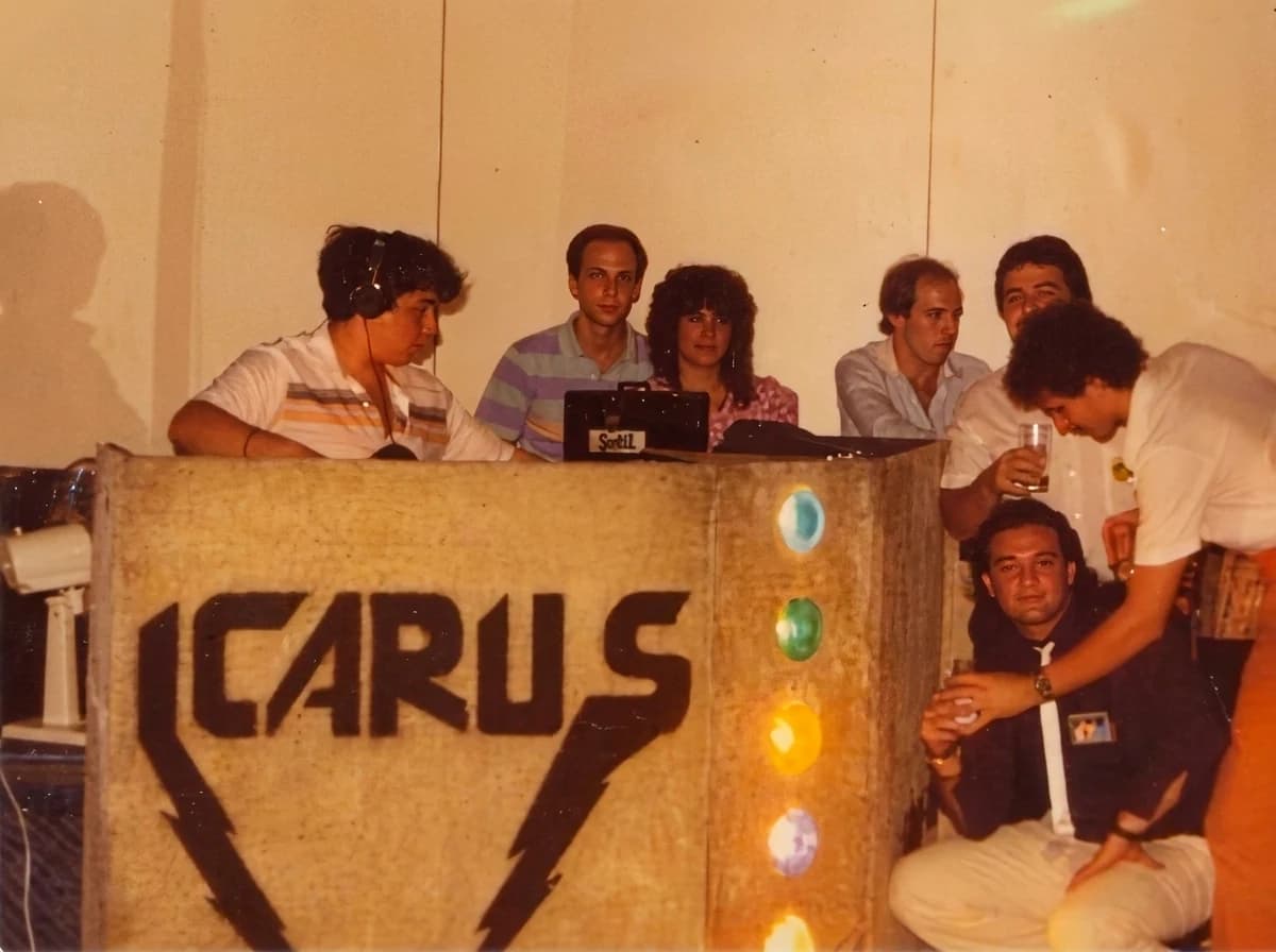 Miniteca Icarus — la cabina rotulada «ICARUS» con un motivo de rayos, rodeada por el equipo de discjockeys y acompañantes (fotografía de archivo restaurada digitalmente) Miniteca Icarus — la cabina rotulada «ICARUS» con un motivo de rayos, rodeada por el equipo de discjockeys y acompañantes (fotografía de archivo restaurada digitalmente)