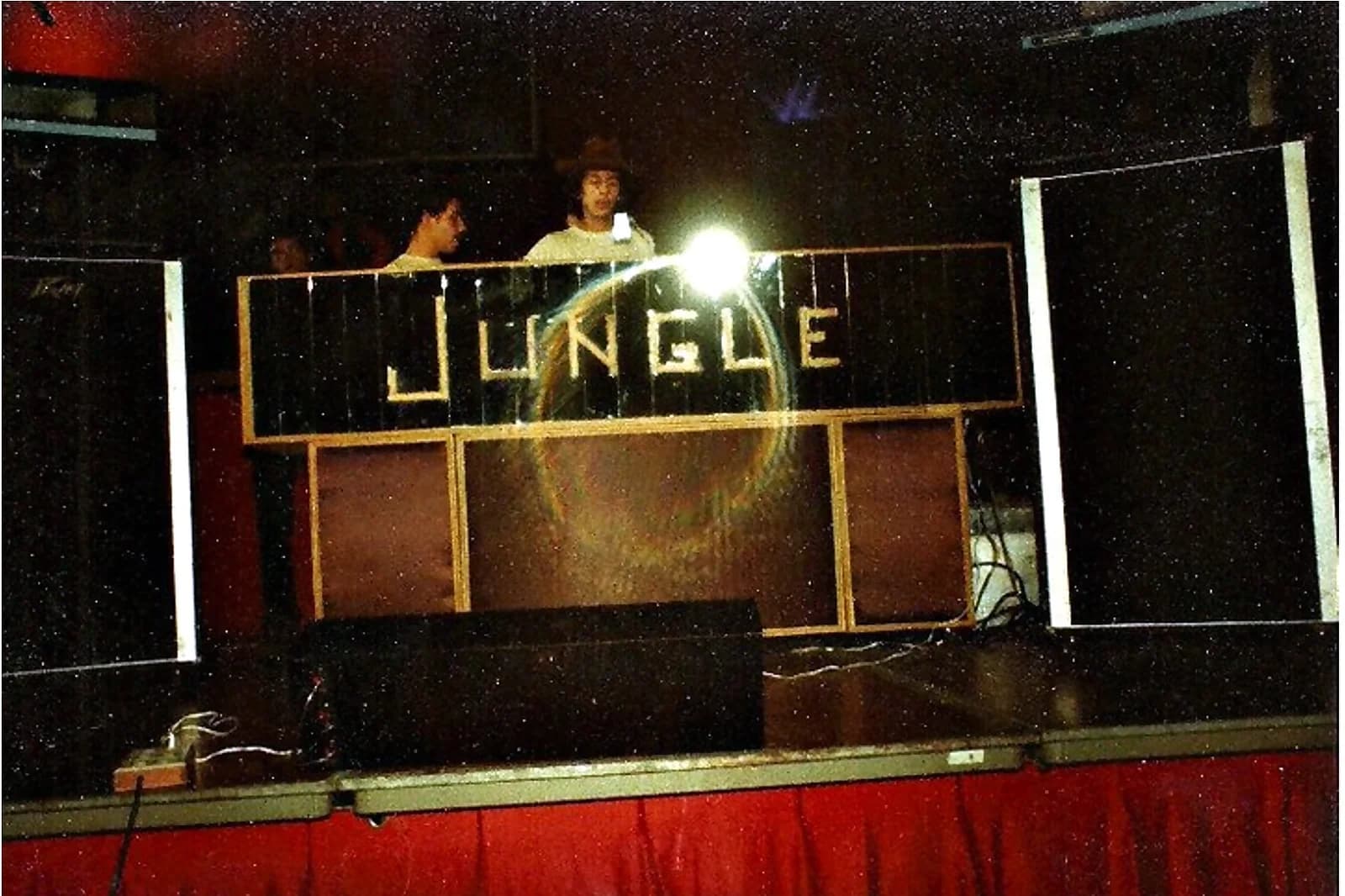 Miniteca Jungle — fotografía de archivo de su cabina con el letrero «JUNGLE» en letras doradas sobre paneles espejados, montada en tarima con dos de sus integrantes tras la consola Miniteca Jungle — fotografía de archivo de su cabina con el letrero «JUNGLE» en letras doradas sobre paneles espejados, montada en tarima con dos de sus integrantes tras la consola