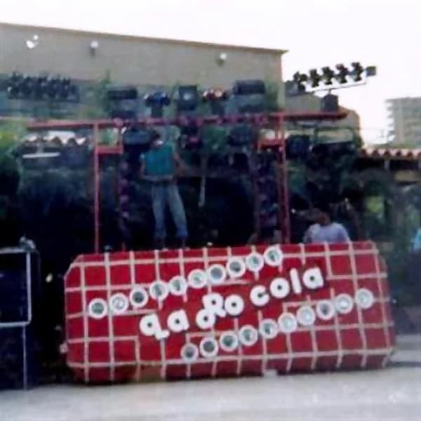 Miniteca La Rocola — el gran cabinón rojo rotulado «La Rocola» con bombillos alrededor, las torres de bafles y un integrante de pie sobre la estructura, en un montaje al aire libre Miniteca La Rocola — el gran cabinón rojo rotulado «La Rocola» con bombillos alrededor, las torres de bafles y un integrante de pie sobre la estructura, en un montaje al aire libre