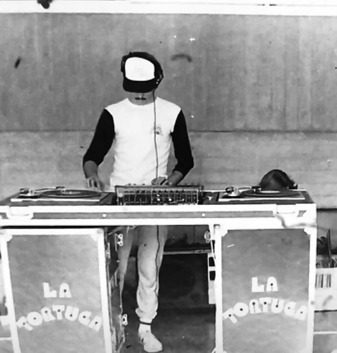 Miniteca La Tortuga — the discjockey behind the booth in a cap and headphones, between two turntables and the mixer; the front panels of the flight case are lettered “LA TORTUGA” Miniteca La Tortuga — the discjockey behind the booth in a cap and headphones, between two turntables and the mixer; the front panels of the flight case are lettered “LA TORTUGA”