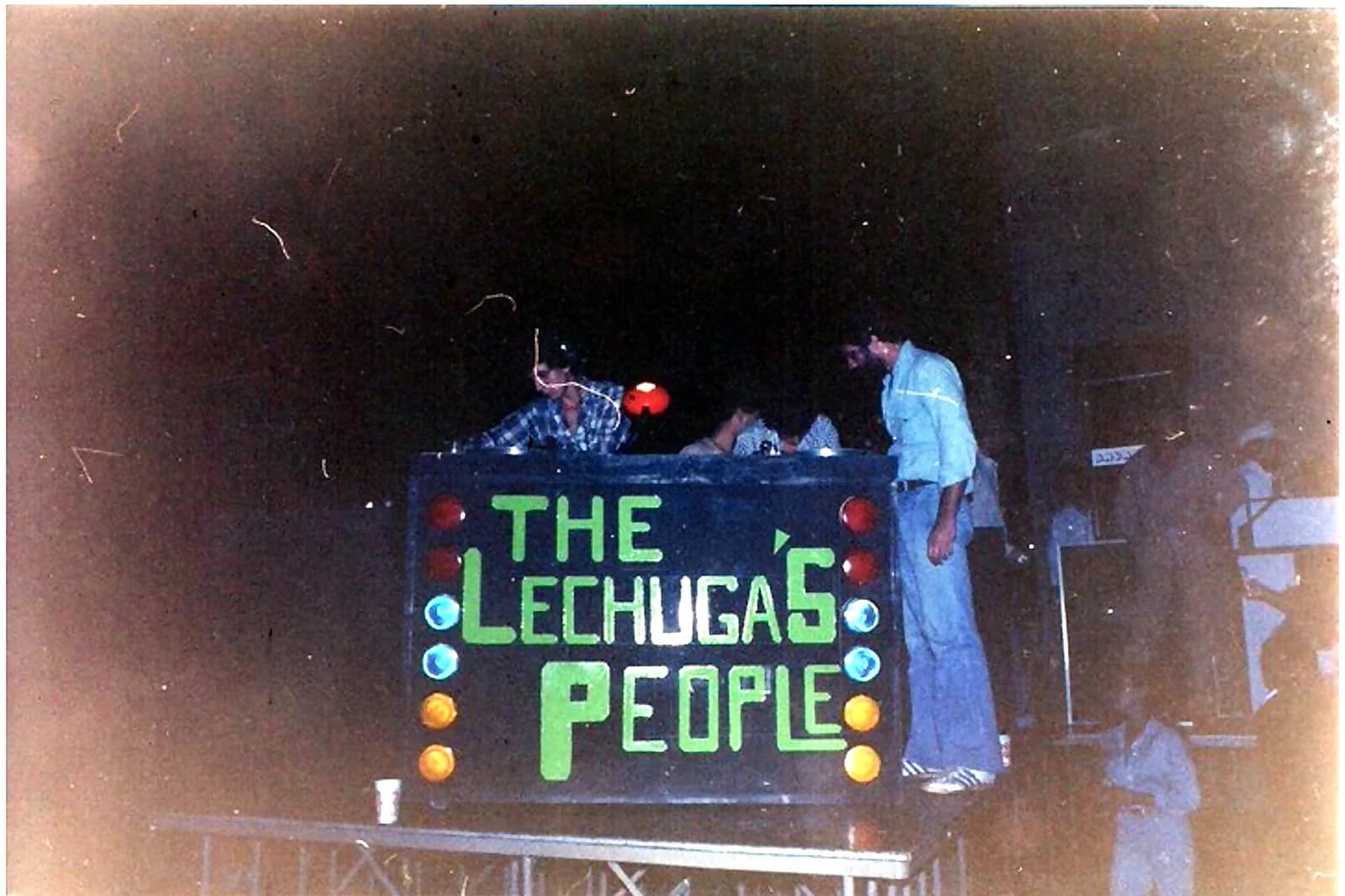 Miniteca The Lechuga's People — la consola con el rótulo verde y filas de luces de colores, con el equipo trabajando sobre la tarima Miniteca The Lechuga's People — la consola con el rótulo verde y filas de luces de colores, con el equipo trabajando sobre la tarima