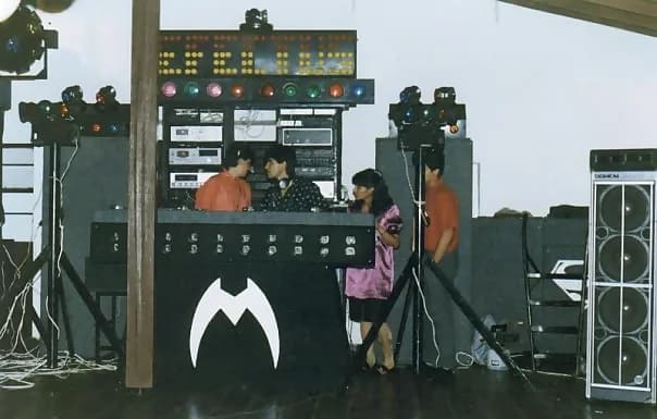 Miniteca Macro Efectus — the booth with the stylized white “M” emblem, the equipment racks, the bars of colored lights and the crew of discjockeys mid-performance (Colombia) Miniteca Macro Efectus — the booth with the stylized white “M” emblem, the equipment racks, the bars of colored lights and the crew of discjockeys mid-performance (Colombia)
