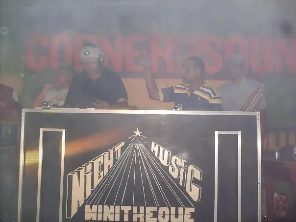 Miniteca Night Music Minitheque — la cabina rotulada «NIGHT MUSIC MINITHEQUE», con una estrella y líneas en fuga, y cuatro integrantes del equipo detrás frente a un telón «Corner Sound» Miniteca Night Music Minitheque — la cabina rotulada «NIGHT MUSIC MINITHEQUE», con una estrella y líneas en fuga, y cuatro integrantes del equipo detrás frente a un telón «Corner Sound»