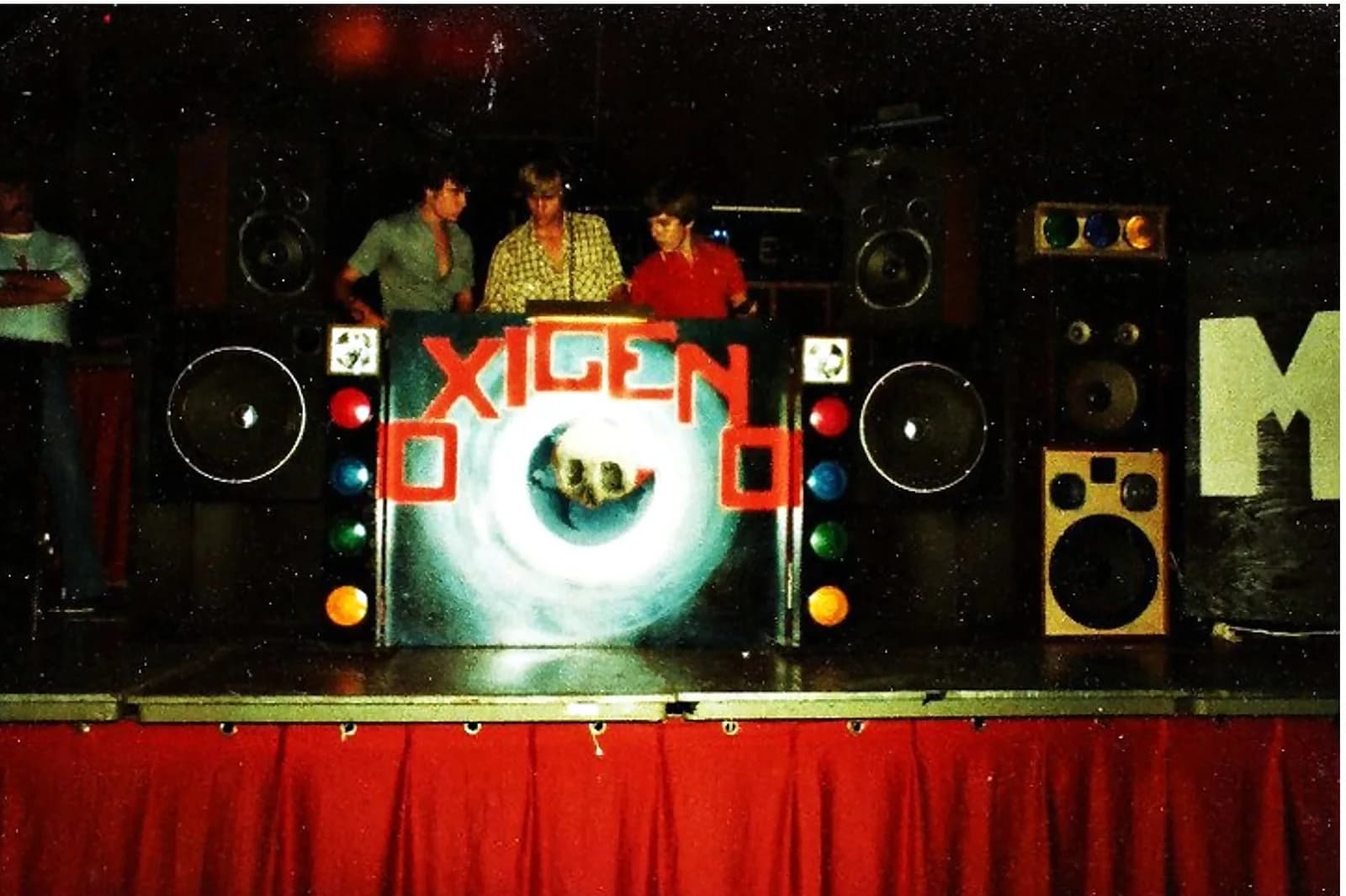 Miniteca Oxígeno — archive photograph of its booth lettered “OXIGENO” in red, with an airbrushed skull at the center, towers of speakers and colored lights on either side and its operators behind the console Miniteca Oxígeno — archive photograph of its booth lettered “OXIGENO” in red, with an airbrushed skull at the center, towers of speakers and colored lights on either side and its operators behind the console