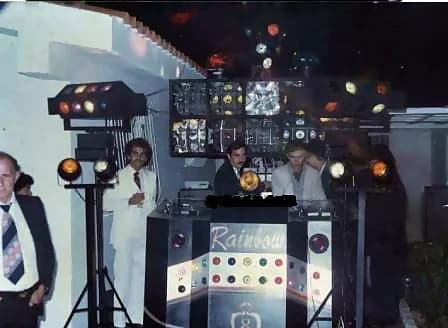 Miniteca The Rainbow — la cabina con el nombre «Rainbow» en el mueble, flanqueada por bancos de luces de colores en una fiesta nocturna; el archivo identifica a Franco Marcone (dueño, de blanco) y Pedro Primera (discjockey) al control Miniteca The Rainbow — la cabina con el nombre «Rainbow» en el mueble, flanqueada por bancos de luces de colores en una fiesta nocturna; el archivo identifica a Franco Marcone (dueño, de blanco) y Pedro Primera (discjockey) al control