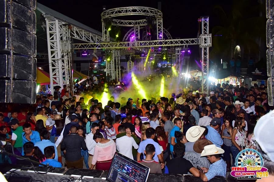 Noche de feria en un recinto ferial: el público llenando la explanada frente a una tarima con truss, cañones de luz y humo, entre las torres de sonido y las carpas de los puestos Noche de feria en un recinto ferial: el público llenando la explanada frente a una tarima con truss, cañones de luz y humo, entre las torres de sonido y las carpas de los puestos
